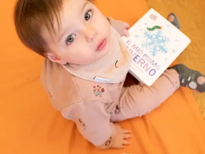 Una bambina piccola, seduta su un tappeto arancione, tiene un libro illustrato