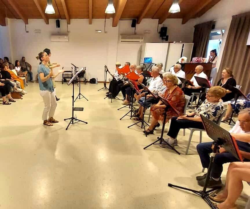 Un coro di persone sedute in cerchio prova in una sala, guidato da una musicista.