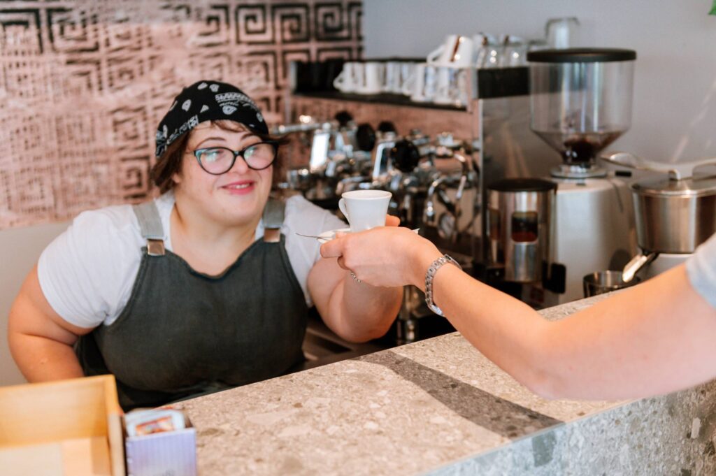 Una giovane barista sorridente porge una tazzina di caffè a un cliente dal bancone.